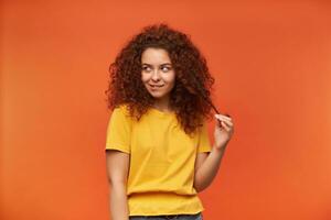 Teenage girl, flirty looking woman with ginger curly hair. Wearing yellow t-shirt. Emotion concept. Play with strand of her hair. Watching to the left at copy space, isolated over orange background photo