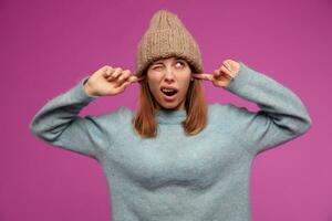 Portrait of young woman with brunette long hair. Wearing blue sweater and knitted hat. Emotion concept. Close her years, don't want to hear. Watching up at copy space, isolated over purple background photo