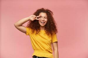 Portrait of attractive, redhead girl with curly hair. Wearing yellow t-shirt. Emotion concept. Showing peace sign over her eye. Watching to the left at copy space, isolated over patel pink background photo