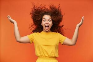 Teenage girl, crazy looking woman with ginger curly hair. Wearing yellow t-shirt. Emotion concept. Lifted her hands up, happy, blow your mind. Watching at the camera isolated over orange background photo