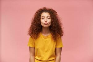 Portrait of attractive, redhead girl with curly hair. Wearing yellow t-shirt. People and emotion concept. Wants to kiss you. Watching at the camera isolated over pastel pink background photo