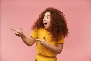 Portrait of attractive, happy redhead girl with curly hair. Wearing yellow t-shirt. People and emotion concept. Watching and pointing to the left at copy space, isolated over patel pink background photo