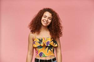 Portrait of attractive, redhead girl with curly hair. Wearing colorful off-shoulder blouse. People and emotion concept. Watching at the camera with cute smile, isolated over pastel pink background photo