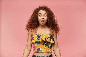 Teenage girl, shocked looking woman with ginger curly hair. Wearing colorful off-shoulder blouse. People and emotion concept. Watching at the camera isolated over pastel pink background photo