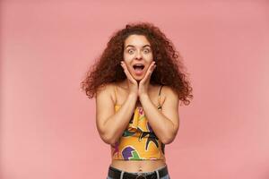 Teenage girl, excited woman with ginger curly hair. Wearing colorful off-shoulder blouse. People and emotion concept. Touching her face. Watching at the camera isolated over pastel pink background photo