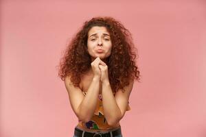 Cute looking woman, beautiful girl with curly ginger hair. Wearing colorful off-shoulder blouse. Fold palms in begging sign, pout her lip. Watching at the camera isolated over pastel pink background photo