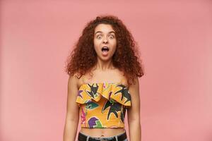 Shocked looking woman, beautiful girl with curly ginger hair. Wearing colorful off-shoulder blouse. People and emotion concept. Watching at the camera isolated over pastel pink background photo