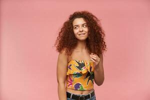 Flirty looking woman with ginger curly hair. Wearing colorful off-shoulder blouse. Playing with strand of hair and biting a lip. Watching to the left at copy space, isolated over patel pink background photo