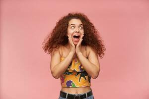 Portrait of excited, redhead girl with curly hair. Wearing colorful off-shoulder blouse. People and emotion concept. Watching to the right at copy space, isolated over pastel pink background photo