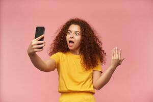 Teenage girl, happy looking woman with ginger curly hair. Wearing yellow t-shirt. Making selfie on a cell phone, looking cool. Asking posture. Stand isolated over pastel pink background photo