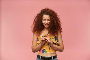 Portrait of attractive, redhead girl with curly hair. Wearing colorful off-shoulder blouse. People and emotion concept. Shyly playing with strand of hair. Stand isolated over pastel pink background photo
