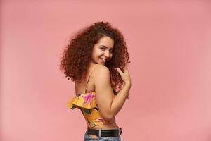 Shy looking woman, beautiful girl with curly ginger hair. Wearing colorful off-shoulder blouse. People and emotion concept. Watching at the camera over shoulder, isolated over pastel pink background photo