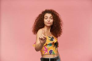 Portrait of refusing, redhead girl with curly hair. Wearing colorful off-shoulder blouse. Emotion concept. Showing finger, reject sign. Watching at the camera isolated over pastel pink background photo