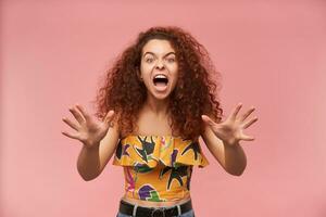 Portrait of angry, redhead girl with curly hair. Wearing colorful off-shoulder blouse. People and emotion concept. Pissed off, had enough. Watching at the camera isolated over pastel pink background photo