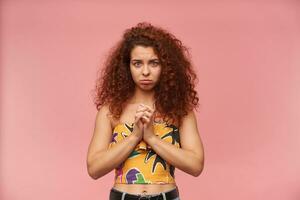 Portrait of upset, redhead girl with curly hair. Wearing colorful off-shoulder blouse. Emotion concept. Fold palms together and ask you. Watching at the camera isolated over pastel pink background photo