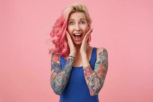 Portrait of young happy amazed nice pink haired lady, with palms on cheeks, her boyfriend proposed to her, looks at the camera with wide open mouth, feels happiness, stands over pink background. photo