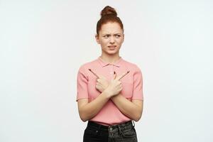 Unhappy lady, frowning woman with ginger hair bun. Wearing pink t-shirt and black jeans. Watching confused to the right and pointing both sides at copy space, isolated over white background photo