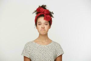Dissatisfied young female pursed lower lip, being abused by something unpleasant, has unhappy expression, dressed t shirt with black polka dots, stands indoor against white wall photo
