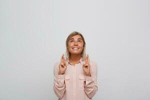 Portrait of happy excited blonde young woman with braces on teeth wears pink shirt keeps fingers crossed, looks up and makes a wish isolated over white background photo