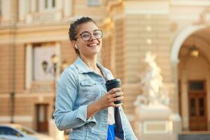 Pretty blonde young girl in glasses walks around the city in a denim jacket, drinks coffee, listens in headphones to favorite music, smiles to the camera photo