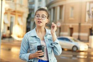 Blonde young girl in glasses walks around the city dressed casually, drinks coffee, listens in headphones to favorite music, took one earphone out of ear and turned around at the call of a passerby. photo