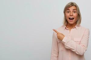 Closeup of happy amazed blonde young woman with braces on teeth and opened mouth wears pink shirt looks surprised and points to the side with finger isolated over white background photo