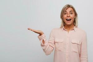 Portrait of smiling amazed blonde young woman with braces on teeth and opened mouth wears pink shirt looks surprised and holding copyspace on palm isolated over white background photo