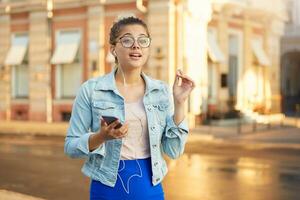 Outdoor shoot of young girl in glasses walks around the city dressed casually, listens in headphones to favorite music, took one earphone out of ear at the call of a passerby or ask for directions. photo