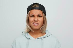 Closeup of sad unhappy blonde young woman with braces on teeth wears black cap and hoodie looks upset and displeased isolated over white background photo