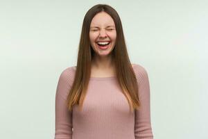 Young woman laughs at a good joke, looks funny, amusing, cheerful, closes eyes with happiness and toothy smile, dressed in a tight pink knit sweater, isolated over white background photo