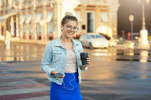 Shiny sunny outdoor photo of pretty blonde young girl in glasses walks around the city in a denim jacket and blue skirt, drinks coffee, listens in headphones to favorite music, smiles to the camera