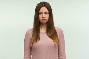 Offended young female feels insult, being abused by something unpleasant, has unhappy expression, dressed in casual pink knit sweater, stands indoor against white wall photo