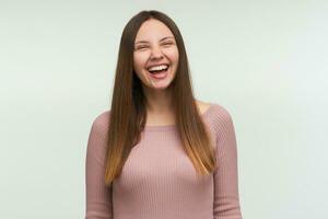 riendo joven mujer con largo pelo abajo, mira divertido, divertido, alegre, se ríe atornillar arriba su ojos, vestido en un apretado rosado tejer suéter, aislado terminado blanco antecedentes foto