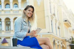 Young girl student after lectures sits, drinks coffee, listens in headphones music, reads something on white tablet, dressed a denim jacket and blue skirt, studying, smiles to the camera photo