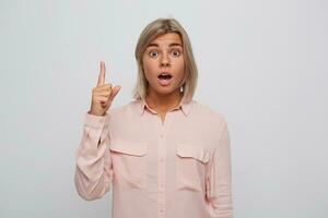Closeup of shocked astonished blonde young woman with opened mouth wears pink shirt looks amazed and points up with finger isolated over white background photo