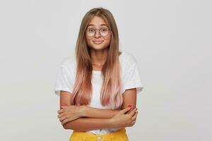 Closeup of shy smiling young woman with long dyed pastel pink hair wears t shirt and spectacles feels embarressed and keeps hands folded isolated over white background photo