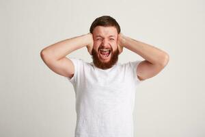 Portrait of mad crazy bearded young man wears t shirt covered ears by hands, keeps eyes closed, feels angry and shouting isolated over white background photo