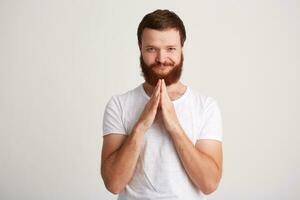 Closeup of happy handsome young man with beard wears t shirt standing with hands folded, touching his chin and thinking isolated over white background photo