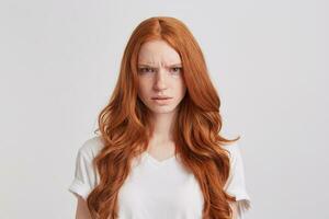 retrato de enojado ceñudo joven mujer con largo ondulado rojo pelo usa t camisa mira enojado y irritado aislado terminado blanco antecedentes foto