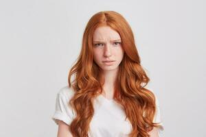 Closeup of unhappy worried redhead young woman with long wavy hair wears t shirt feels angry and irritated isolated over white background photo