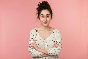 Self-confident young dark-haired curly girl standing with crossed arms, looking with interest and curiosity, dressed in blouse with polka dots, isolated on pink wall photo