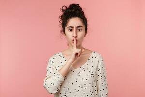 lose up of concerned worried beautiful dark haired girl with making silence gesture putting fore finger on the lips, asks to stay quiet, keep secret, isolated over pink background photo