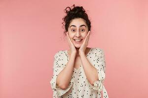 Astonished delighted, recognized something unusual, new, young dark-haired curly girl keeps palms near the face, in shirt with polka dots, on her face an expression of amazement, isolated on pink wall photo