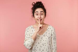 lose up of happy excited beautiful brunette girl making silence gesture putting fore finger on the lips, asks to stay quiet, keep silence, secret, isolated over pink background photo