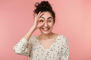 Indoor portrait of funny cute attractive brunette girl with collected wave hair making okey symbol close to eye with one hand, widely smiling, in blouse with polka dots,isolated on pink background photo