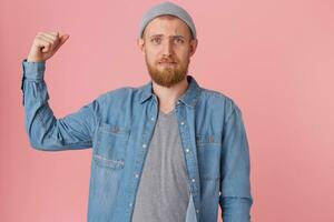 A young bearded guy can not boast of his strength, displeased with his physical form, raised his bent arm to demonstrate weak muscles, with chagrin looking at the camera, dressed in a denim shirt photo