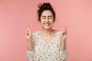 Studio shot of young female keeping her fingers crossed and eyes closed while making a wish. Charming, positive girl sincerely believing in her luck, hopes for the best, isolated on pink background photo