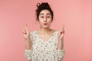 Charming young female keeping her fingers crossed and eyes widely opened sending air kiss, making a wish or thanks someone for the favor. Girl hopes for the best, isolated on pink background photo