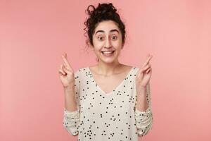 emocionado joven hembra acuerdo su dedos cruzado y ojos extensamente abrió mientras haciendo un desear. encantador, positivo niña atentamente creyendo en su suerte, esperanzas para el mejor, aislado en rosado antecedentes foto