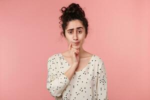 Attentive young woman looks with suspicion standing holds hand near chin and frowning, with thoughtful suspicious mistrustful look, doubts, has an incredulous look, isolated over pink background photo